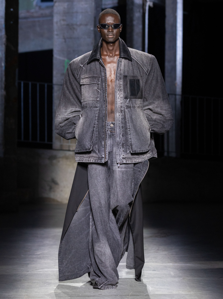 Model walks the runway in boxy Juun.J denim work jacket and wide-leg denim jeans with additional draping on the leg with hands in their pockets at Paris Fashion Week