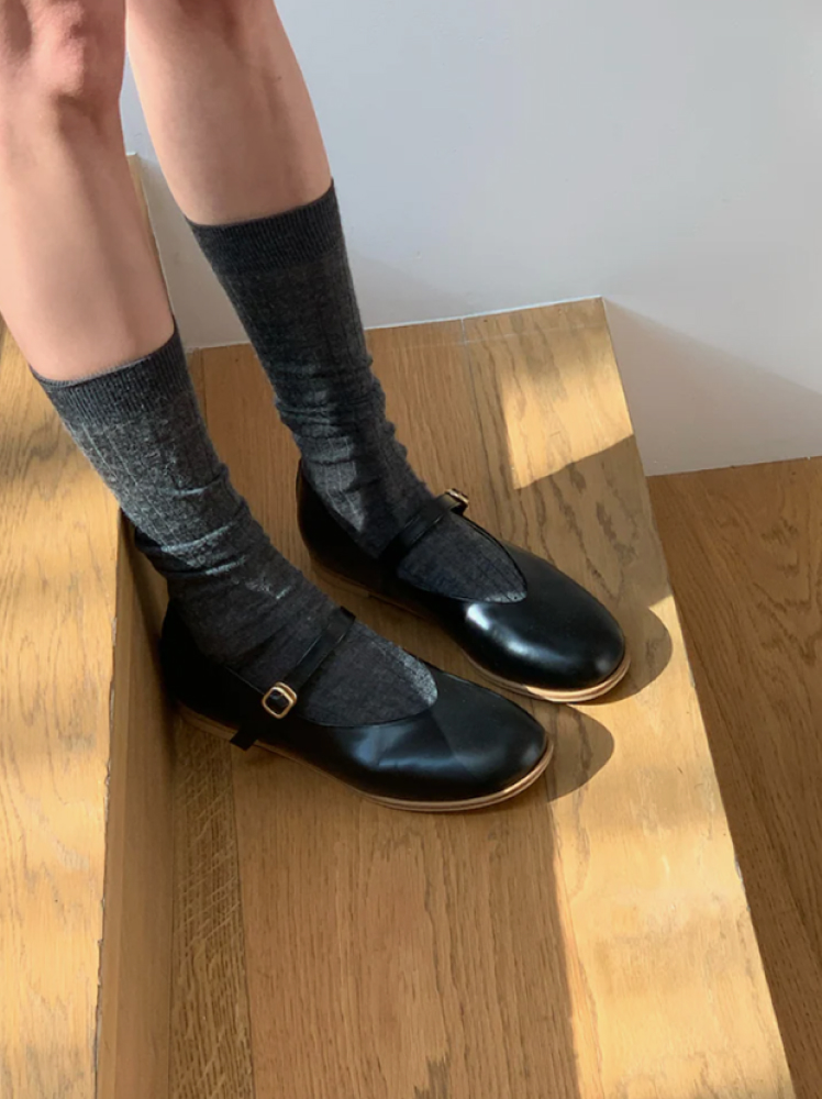 A woman stands on a wooden step in the sun while wearing black leather mary jane shoes by the brand Beanpole, a label owned by the Samsung C&T Fashion Group, and she has paired the shoes with a pair of dark grey socks that go up to her mid-calf.