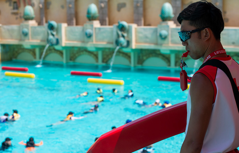 A lifeguard at Everland’s Caribbean Bay holds his whistle in his mouth and looks on at guests swimming in life jackets in the water park’s outdoor wave pool.