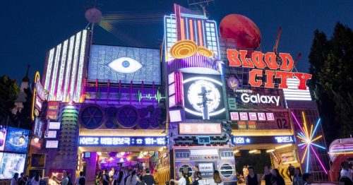 Neon-lit exterior of Everland's "Blood City" horror-themed zone in collaboration with Netflix at night, featuring colorful signs and decorations, with visitors walking nearby.