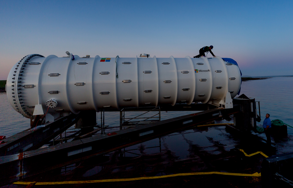 An underwater data center sits next to the sea as the sun sets in the background, ready to be submerged, with a worker situated on top of the data center doing final checks.