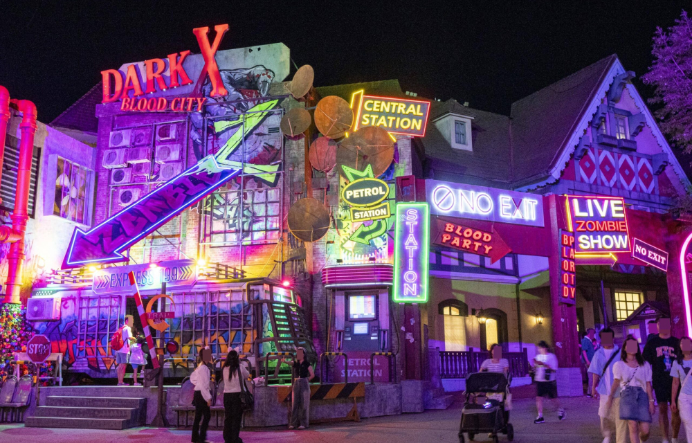 Neon-lit exterior of Everland's "Blood City" horror-themed zone at night, featuring colorful signs and decorations, with visitors walking nearby.