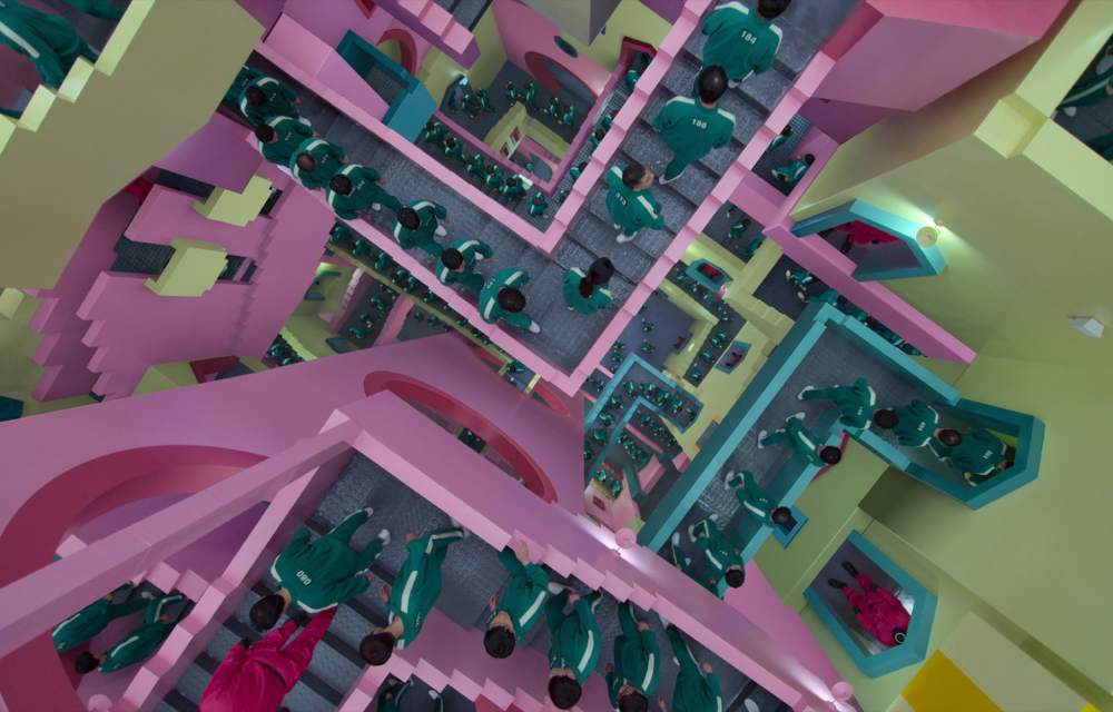 A surreal, maze-like staircase scene from "Squid Game," featuring multiple players in green tracksuits navigating the pastel-colored, Escher-inspired environment.