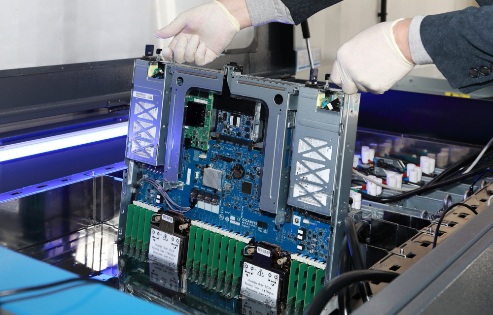 A worker holds up a component of a server that is immersed in a coolant that keeps it cool with printed circuit boards and cables on display.