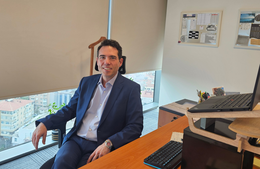 A businessman in a suit sits in front of a computer on his desk in a desk chair, placed in front of a window with a view of the city peeking out below the blinds.