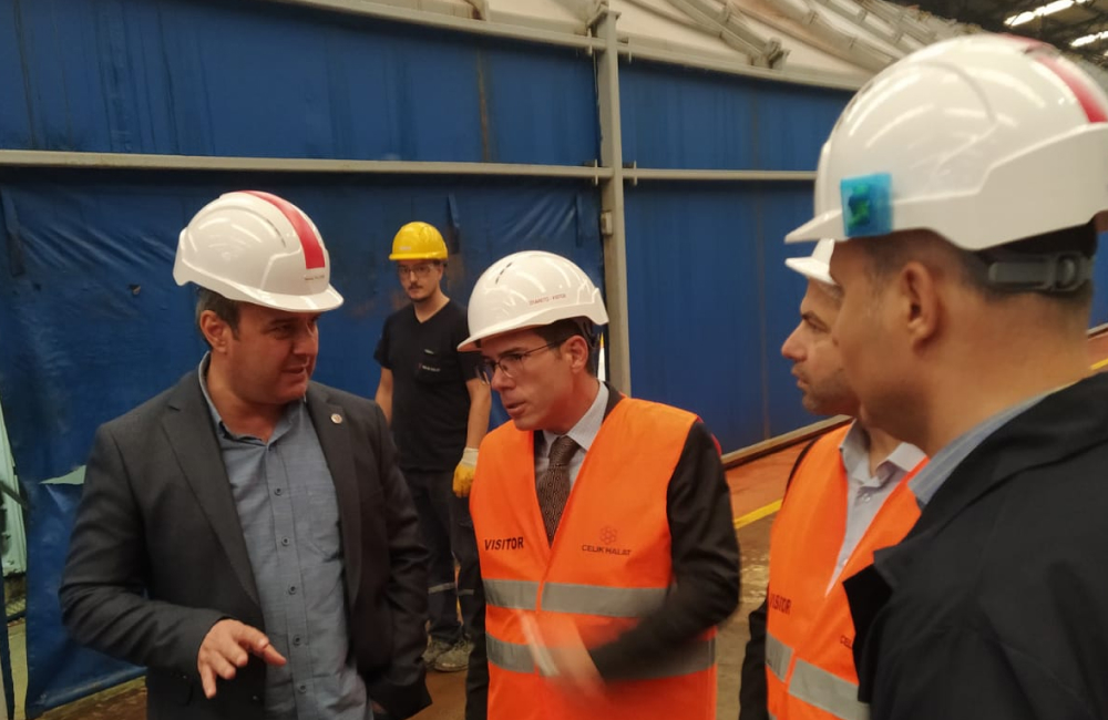 Four businessmen in hardhats and high vis vests stand around and discuss business in a factory while wearing suits.