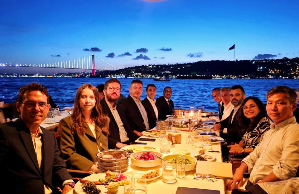 A group of eleven colleagues formally dressed sit around a table at a restaurant that looks out to the ocean to enjoy a meal together.