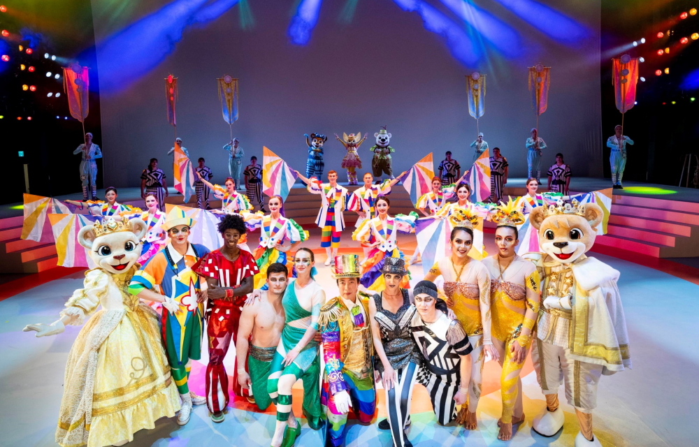 A cast of circus performers pose on their stage in full costumes