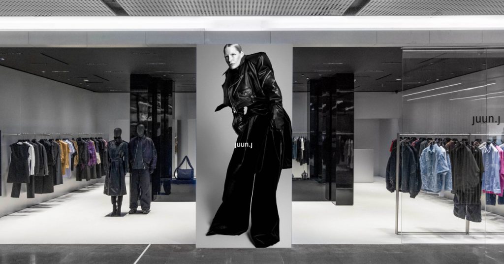 The front entrance to a luxury fashion store with a black glossy interior, bright lights, and black and monotoned clothing items hanging up on display throughout the store.