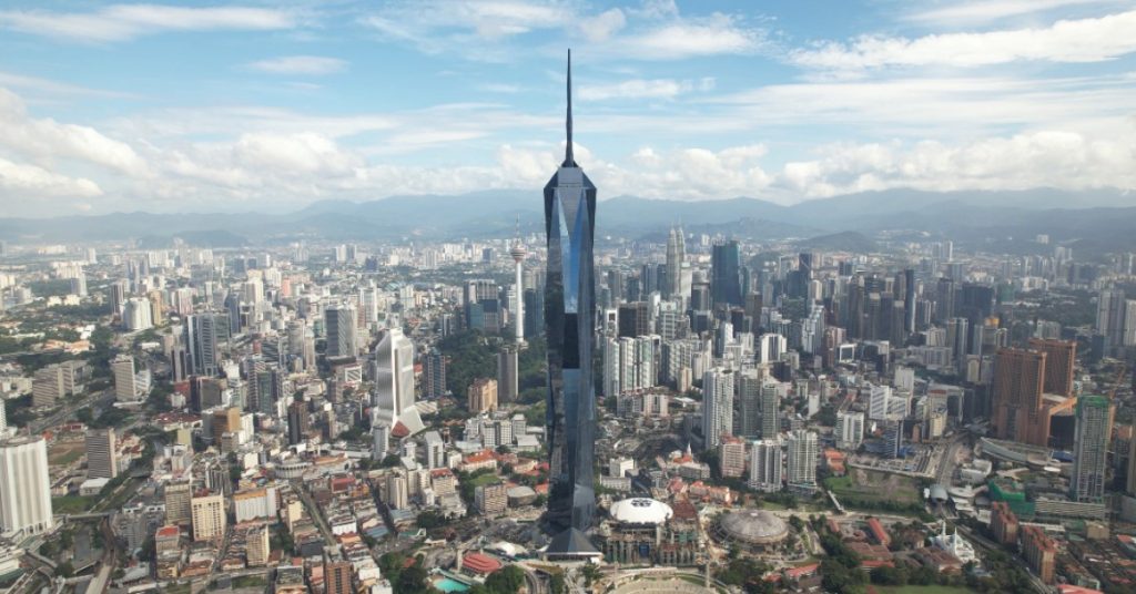 A wide shot of Merdeka 118, the towering diamond-like skyscraper standing tall in Kuala Lumpur surrounded by parks and other high-rise buildings.