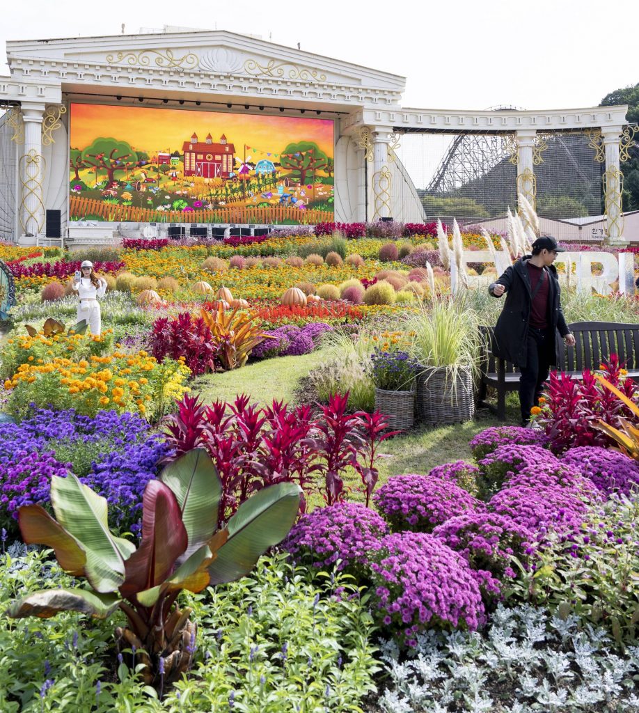 A beautifully manicured garden featuring autumnal plants and flowers in yellow, orange, and purple tones, placed in front of a large stage at the Everland theme park.