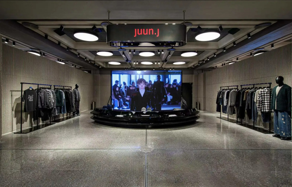 A large fashion retail interior space with concrete flooring, large round lights, a TV screen in the center of the store displaying the fashion brand’s collection, and garment racks lined with dark-colored clothing along either side of the room.