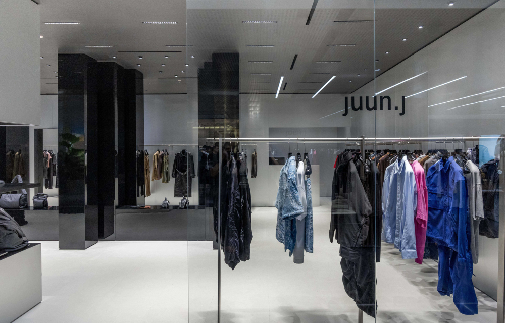 The glass window of Juun.J’s flagship store at REEL Shanghai, with bright blue, pink and black colored clothing on display in a grey store with accents of glossy black paneling.