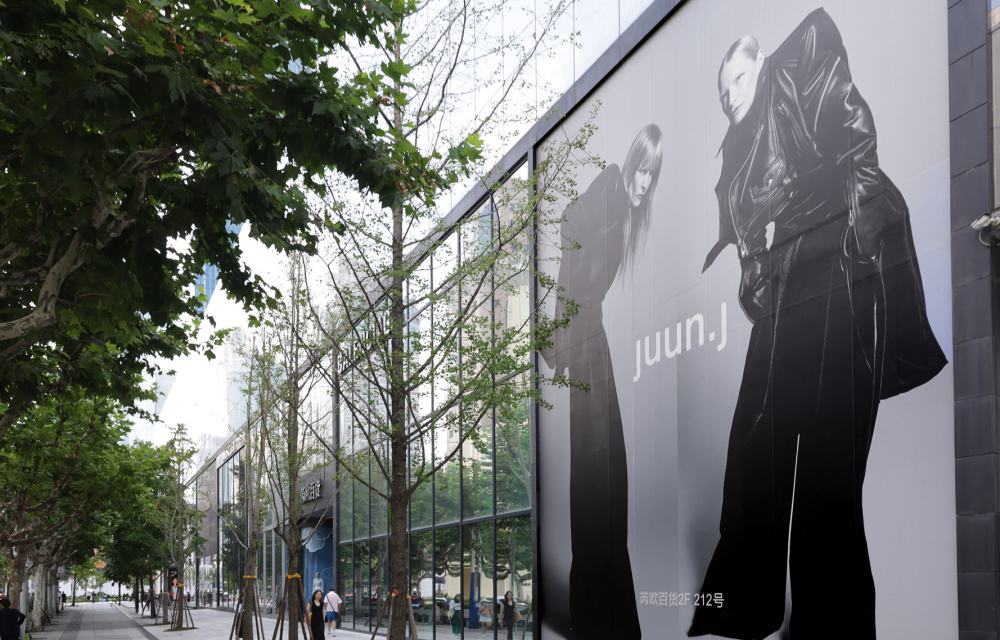 A fashion advertisement featuring models wearing black Juun.J clothing is featured on a building’s exterior alongside glass paneling. The building is located on a tree-lined street with a wide pedestrian footpath.