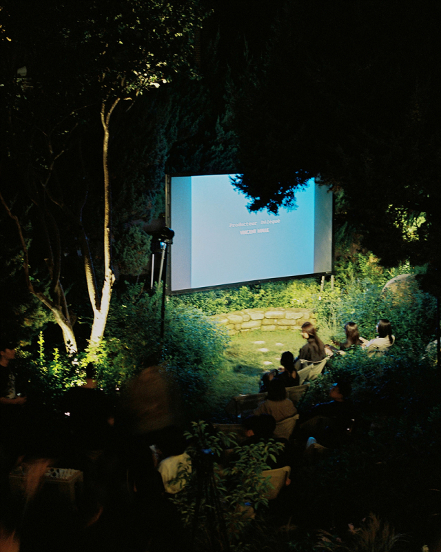 An outdoor movie theater is set up for a film screening, people are seated to watch the film that is being projected on a screen surrounded by trees in a garden.