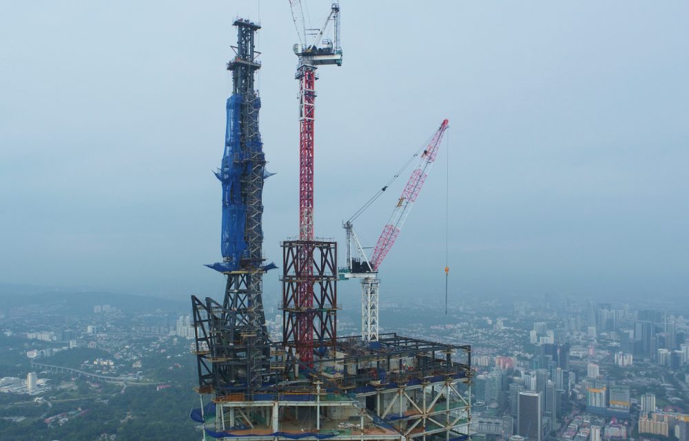 Construction work being done at an extreme height on Merdeka 118 on its 160m spire. Three cranes are pictured at the top of the structure which will become the base of the tower's spire. In the background through a hazy sky is the city of Kuala Lumpur.