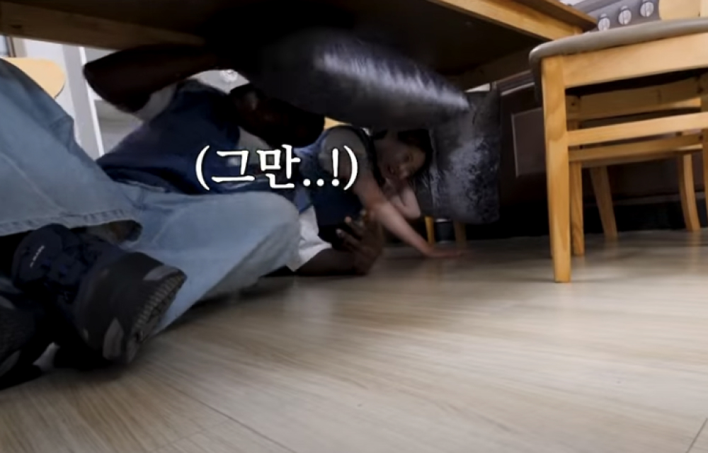 A woman and a man take cover under a wooden table during an earthquake simulation with cushions placed over their heads.