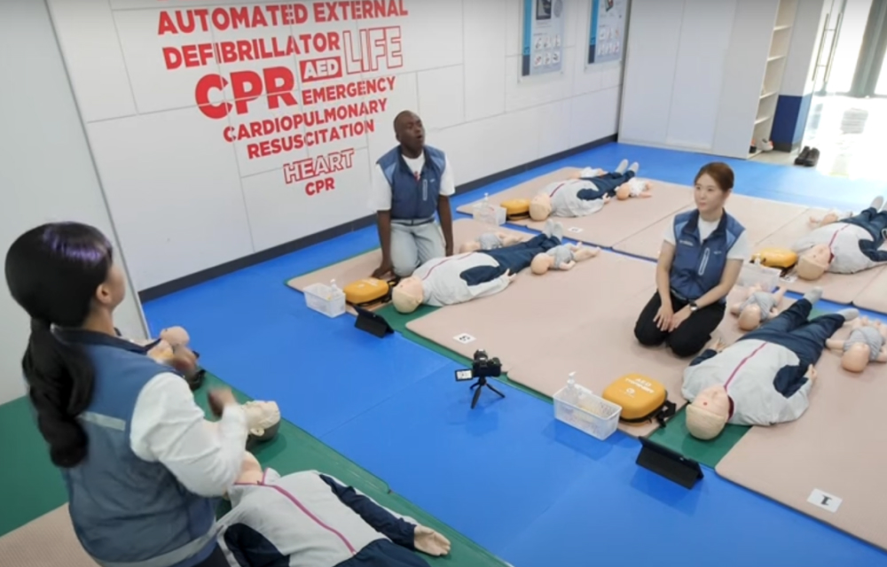 A female instructor educates a man and a woman in a classroom about the basic steps of performing CPR. On the ground lay CPR mannequins on floor mats as part of the experiential education zone at Samsung C&T’s Safety Academy.