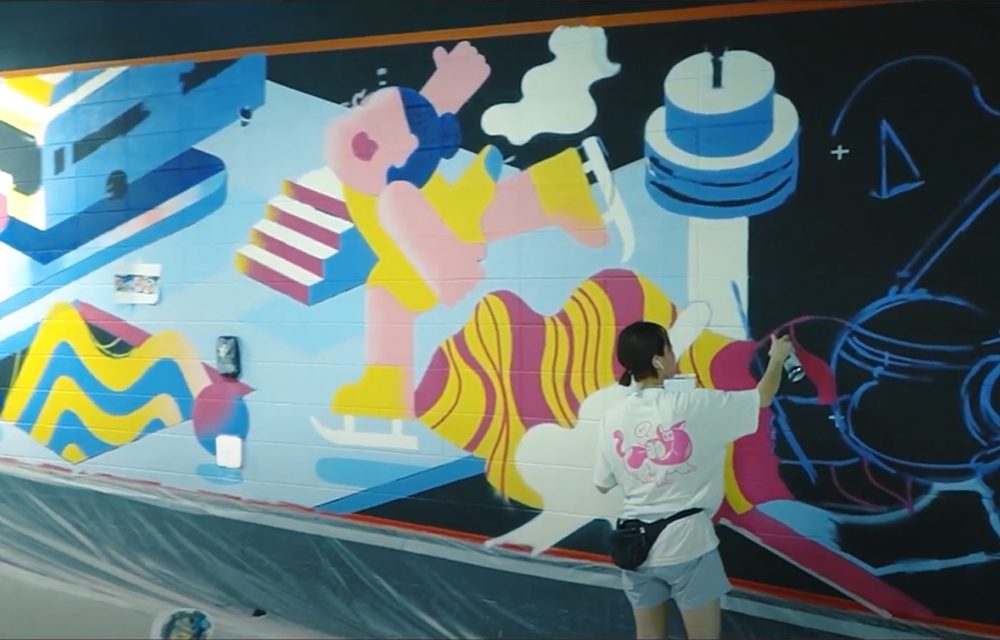 An artist painting a large, colorful mural on a wall, showcasing bold shapes and vibrant colors, wearing a white shirt and standing in front of the artwork.