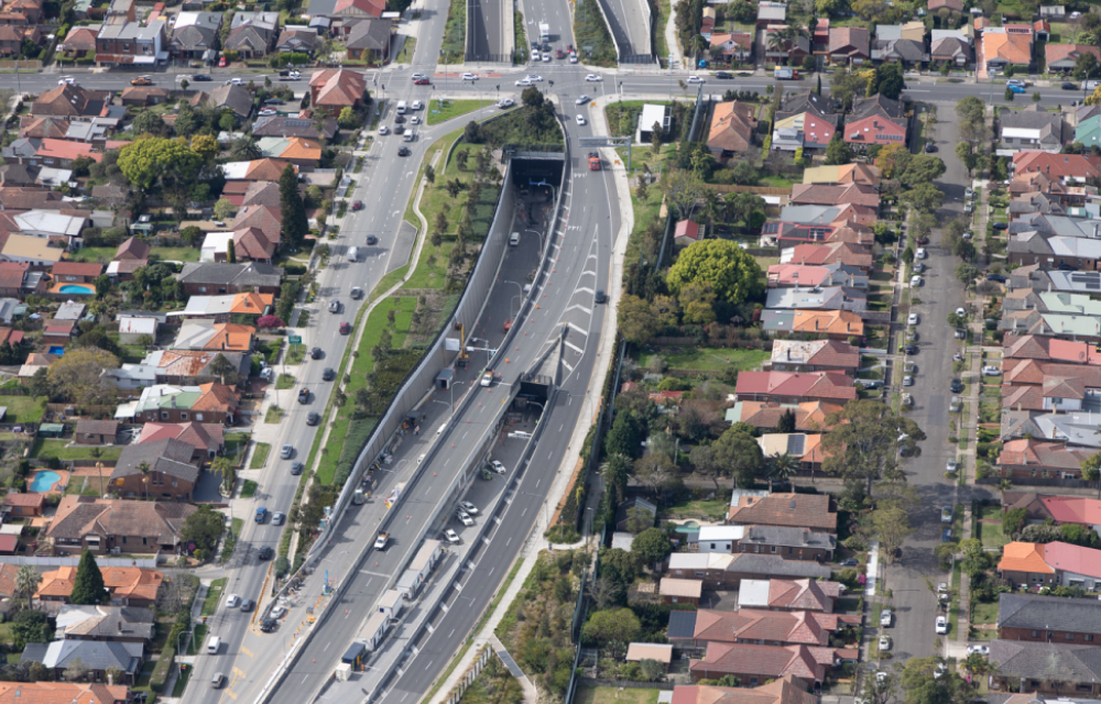 A major highway in Sydney, the WCX3a, runs through the center of a densely populated residential area. The highway leads to an underground tunnel and is surrounded by lush trees and vegetation. The construction was completed by Samsung C&T and other partners.