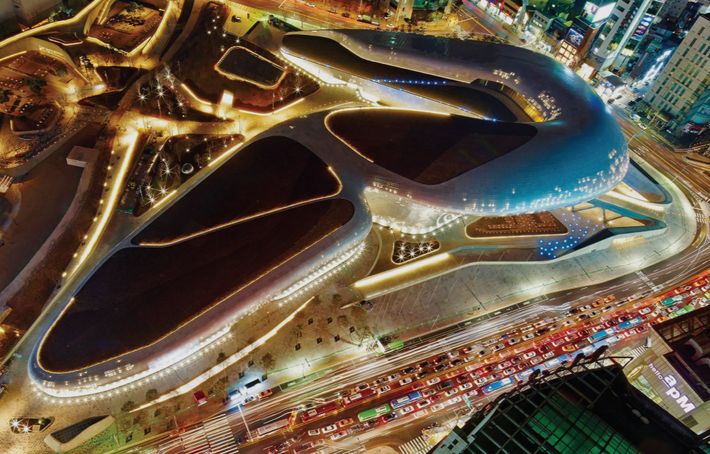 An ariel view of the DDP illuminated at night showcasing its sweeping curves and architectural brilliance, set against the backdrop of bustling streets filled with the vibrant energy of Seoul.