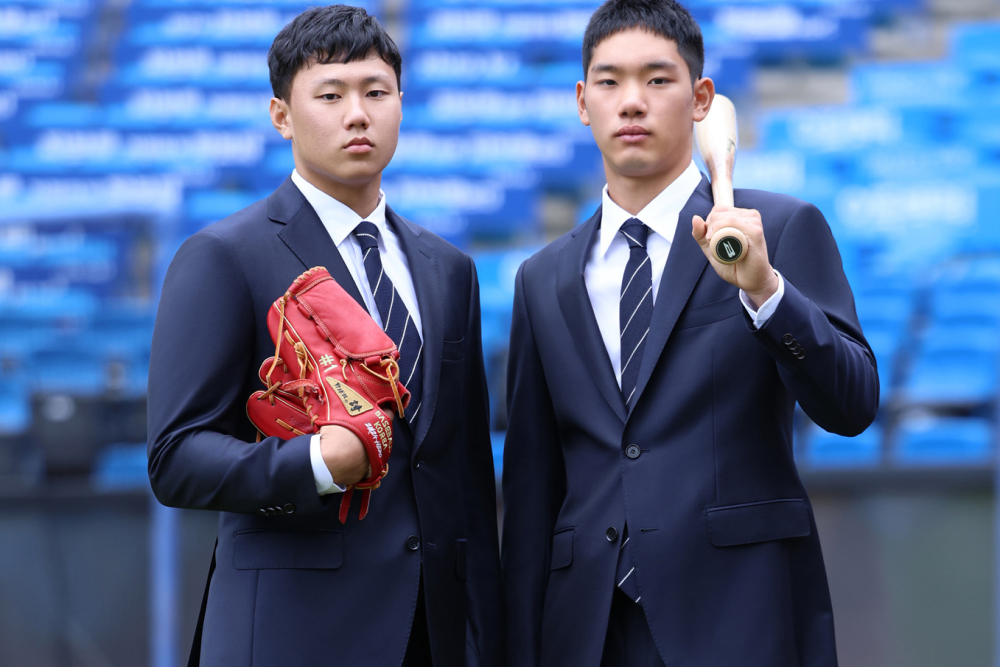 Two rookie baseball players stand side by side in their navy Galaxy GX suits, one player holds a mitt in his hand while the other rests a baseball bat on his shoulder with neutral facial expressions.