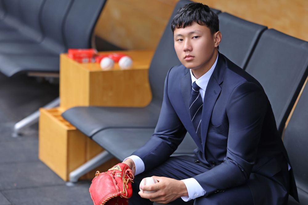 A Korean baseball player poses in a well-tailored navy suit with a baseball glove and a ball in his left and right hands, respectively.