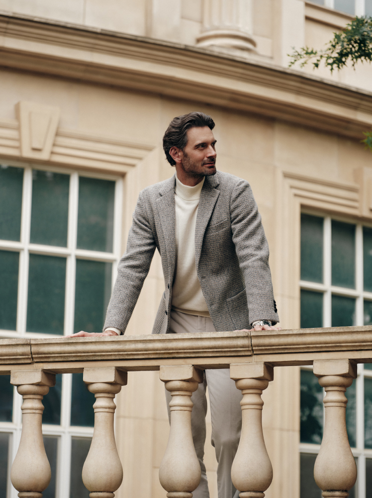 A stylish model stands in front of a traditional style railing wearing a beige wool blazer, white mock neck sweater and relaxed fit trousers.