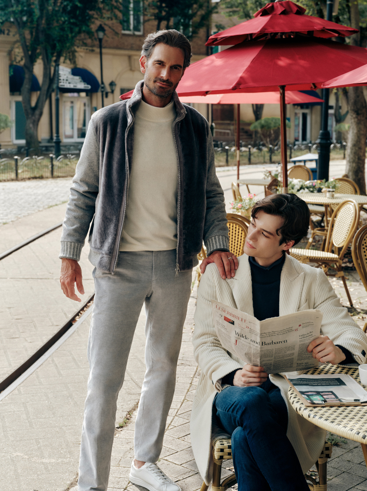 A man dressed in a casual, yet trendy outfit rests his hand on the shoulder of an equally stylish man sitting to read a newspaper.