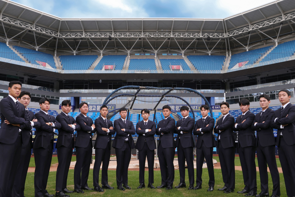 15 baseball players stand in a semicircle formation with their arms crossed over their chests. They are wearing custom Galaxy suits and are standing on a baseball diamond in front of an empty stadium.