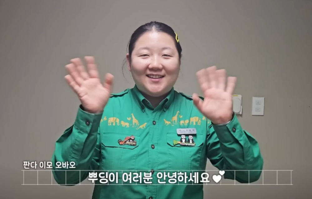 A cheerful panda keeper wearing a green Everland uniform waves cheerfully at the camera during an interview for her YouTube vlog series called ‘Oh with DoongDoong’