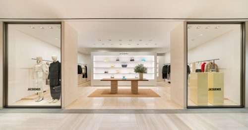 A wide angle image of the new Jacquemus store in Shinsagae, Gangnam, with a wide front entrance and two large glass windows on either side. The center of the store showcases the brand’s signature handbags on a brightly lit display shelf, with a wooden table in the center sitting on top of a rustic raffia rug. In the window on the right hand side sits two pastel yellow plinths that carry the stores symmetrical design.