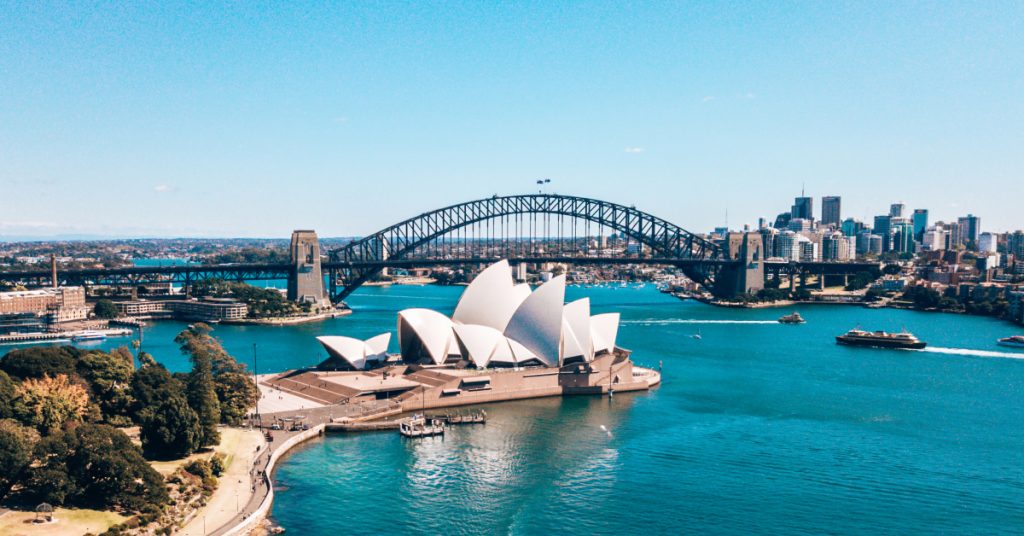 A wide view of Sydney Harbour featuring the Sydney Opera House, the Sydney Harbour Bridge, and ferries in the water commuting passengers back and forth. The water in the harbour is a vivid shade of blue with the sky shining on a beautiful day.