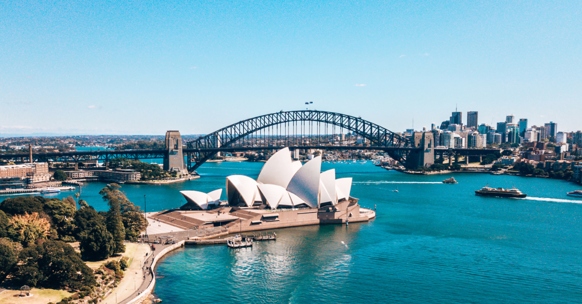 A wide view of Sydney Harbour featuring the Sydney Opera House, the Sydney Harbour Bridge, and ferries in the water commuting passengers back and forth. The water in the harbour is a vivid shade of blue with the sky shining on a beautiful day.