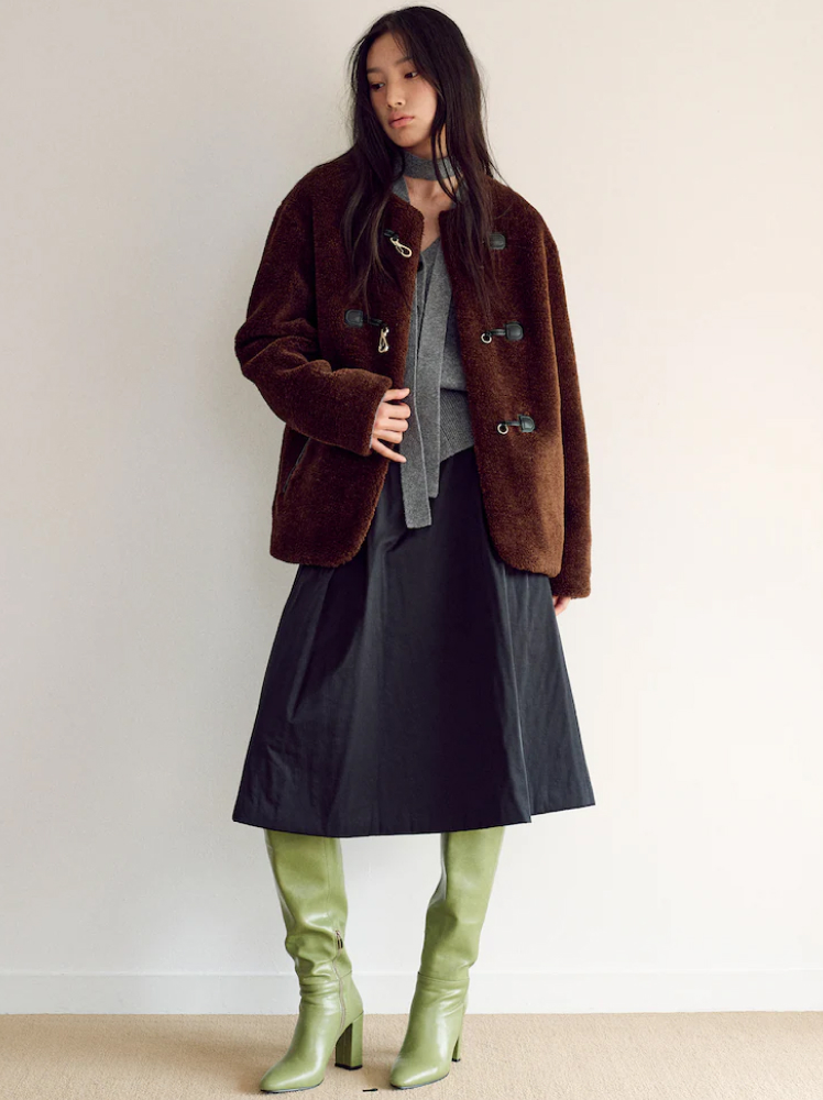 A woman poses in a relaxed stance while wearing a brown fur duffle coat with black and metal toggle fasteners. This is paired with a grey knit cardigan and a thin scarf in the same knit around her neck, an A-line black skirt and a stunning pair of lime green knee-high boots with a tall block heel.