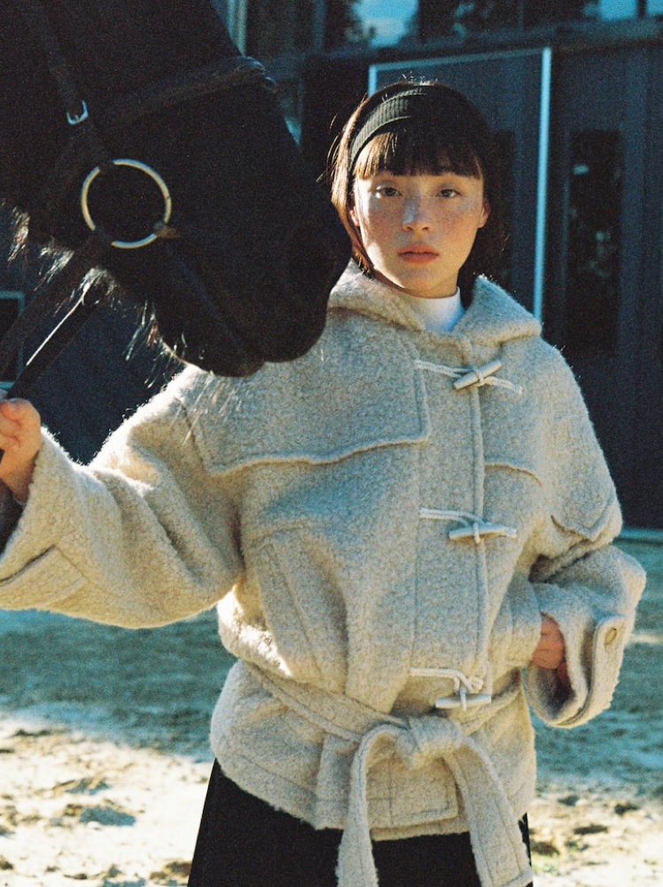 A woman stands beside a horse while holding its reins with her right hand and her left hand resting in the pocket of her fluffy duffle style jacket with a hood and a waist belt detail.