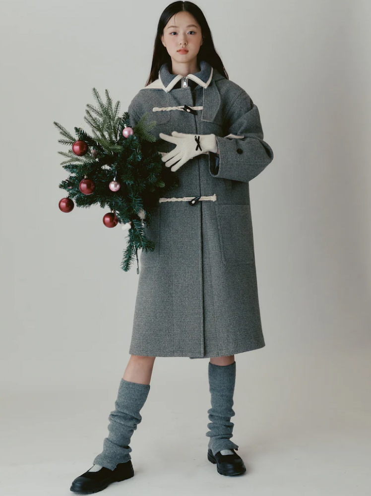 A woman wears a grey duffle coat with white toggle details and the collar from her knit jumper underneath poking through the neckline of the coat. In her hands, the woman holds fragments of an artificial Christmas tree covered in red and pink baubles as though the tree is a floral bouquet. She is also wearing grey leg warmers and chunky black mary jane shoes.