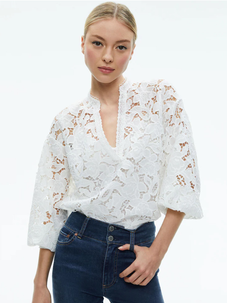 A woman stands with her hand tucked into the belt loop of her high-waisted jeans while wearing a white lace boho-chic style top by Alice and Olivia.