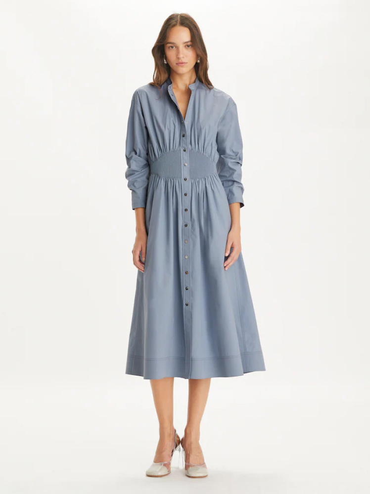 A woman poses in a mid-length gray blue dress with an A-line shape. The dress has long sleeves, a button up collar, and gathered pleats at the waist for a simple, elegant boho-chic look from Tory Burch. The model is wearing heels to complete the look.