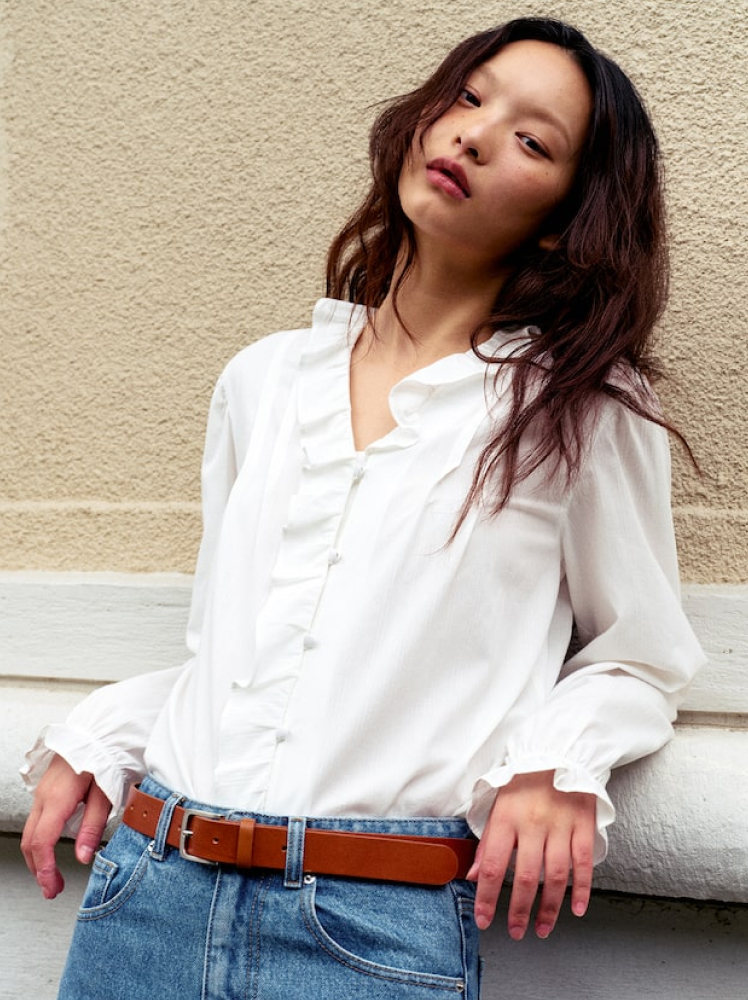 A model leans back against a wall wearing a boho-chic style white blouse with frills along the center, paired with jeans and a tan leather belt.