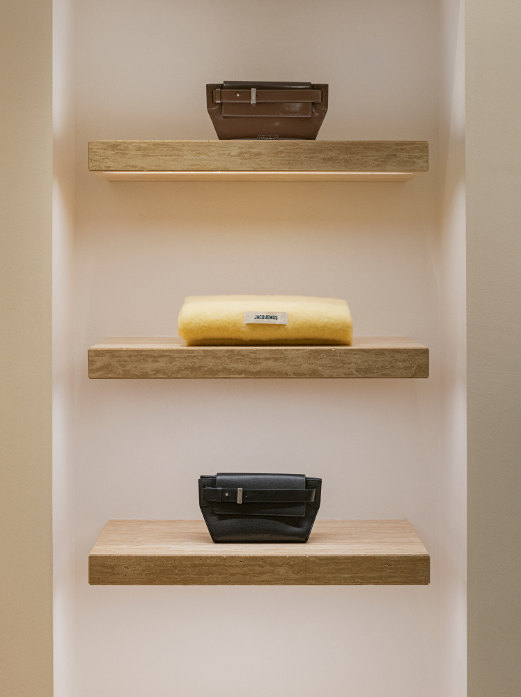 Three floating wooden shelves are lit up, displaying two ‘The Capri messenger bags’ by Jacquemus, one brown and one black. A soft wool scarf sits on the middle shelf, folded neatly with the brand’s label facing outward.