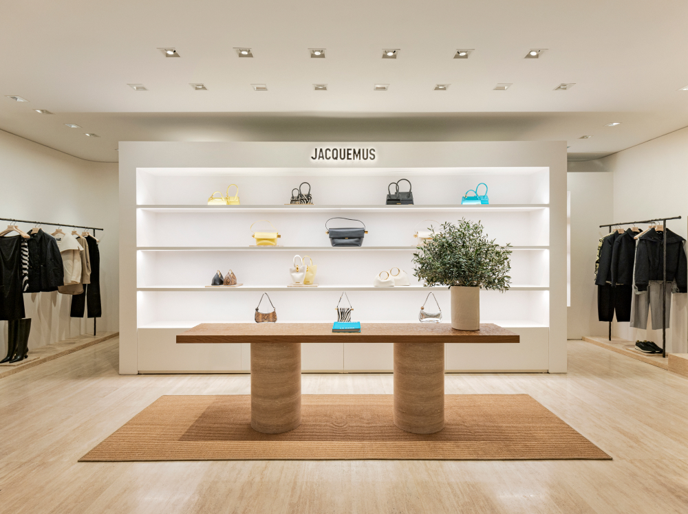 The interior of a Jacquemus store in Gangnam, showcasing the brand’s signature handbags on a brightly lit display shelf, with a wooden table in the center sitting on top of a rustic raffia rug. On either side of the store, clothing racks with the brand’s latest collections are on display.