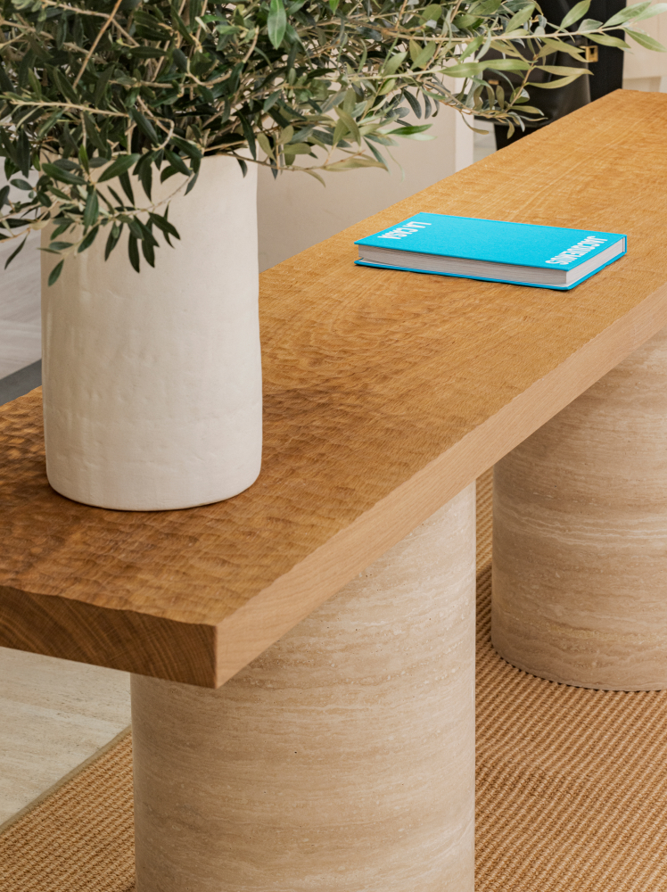 A close up of the store’s centerpiece, a wooden table with two large cylindrical legs. On top of the table sits a plant in a large white vase and a brightly colored blue book.