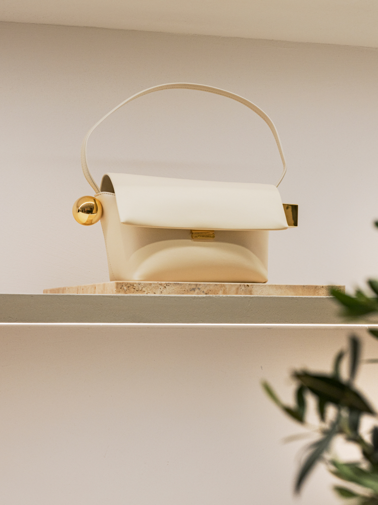 A light ivory colored Jacquemus ‘The Round Carre’ shoulder bag sits on a white shelf at a department store in Seoul. The leather bag has a simple shoulder strap and a flap closure, featuring two gold details, a sphere and a cube, on either side of the bag.