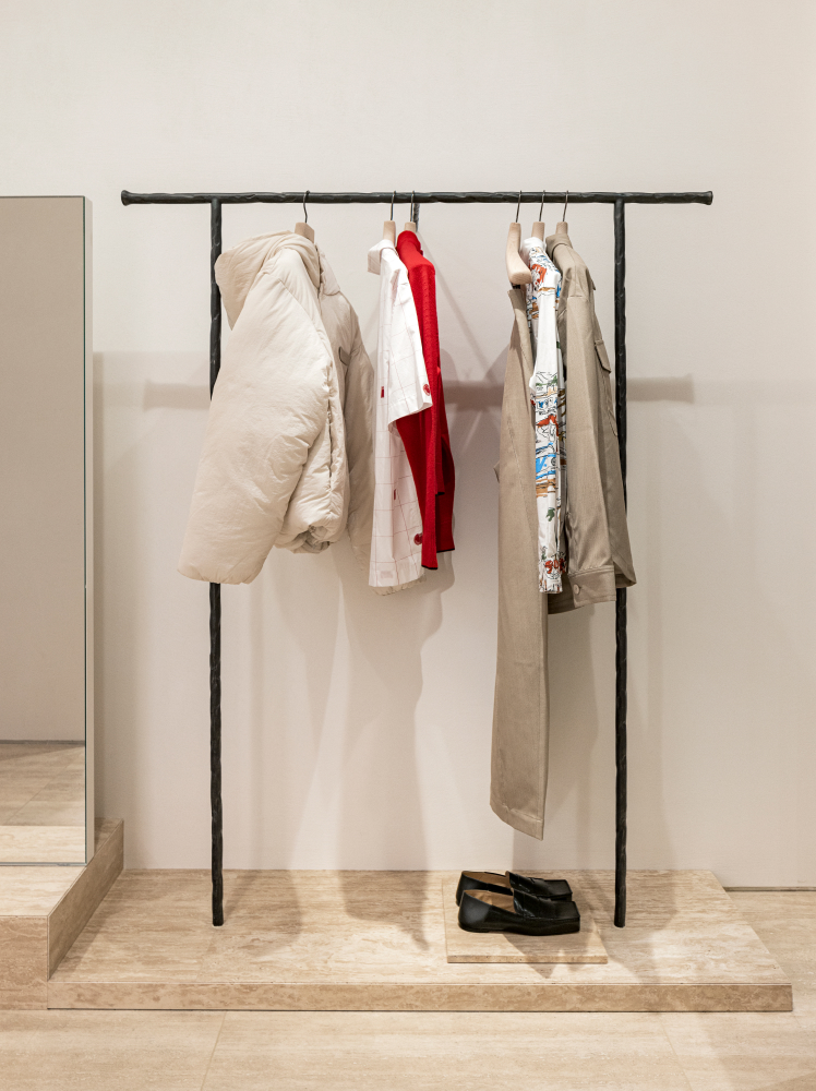 A simple standalone black clothing rack sits on top of white platform where white, red, and cream clothing pieces by French label Jacquemus are hanging on display.