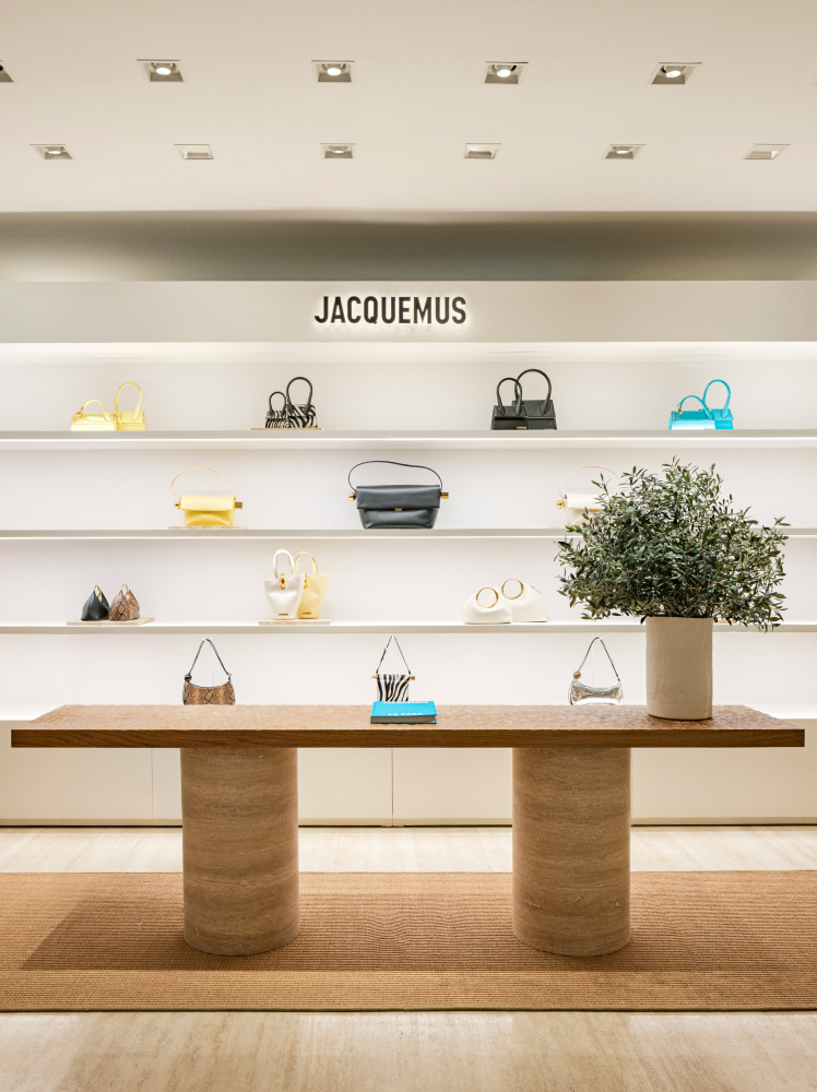 The interior of a Jacquemus store in Gangnam showcases the brand’s signature handbags on a brightly lit display shelf, with a wooden table in the center sitting on top of a rustic raffia rug.