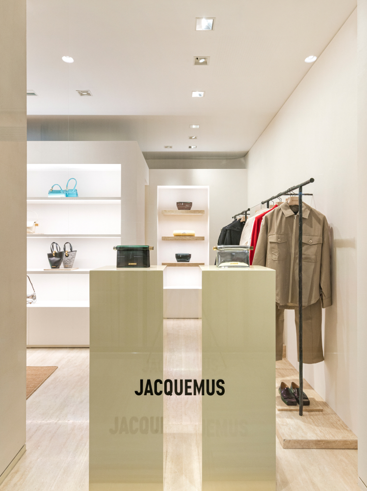 Two pastel yellow plinths stand in the window display of the new Jacquemus Gangnam store with a handbag placed on both. In the background, clothing items from the latest Fall/Winter collections are displayed in a brightly lit interior.