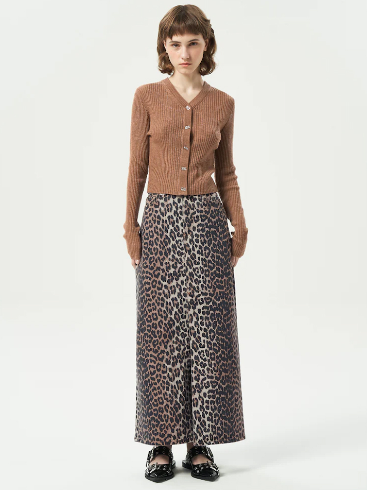 A model poses with her hands by her side wearing a long maxi pencil skirt with a slit down the middle in a leopard print design. The skirt is paired with a buttoned up brown cardigan and black pointed mary jane flats.