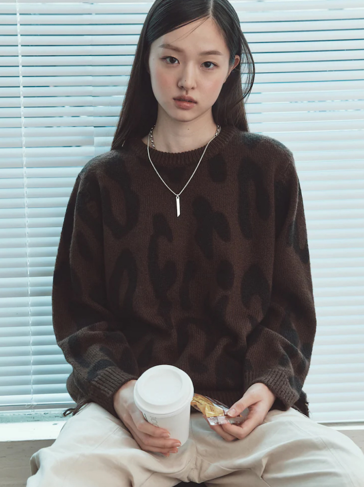 A model sits in front of a window with white venetian blinds wearing a cozy leopard print jumper, white trousers, a simple silver metal necklace and holds a takeaway coffee in her hand.