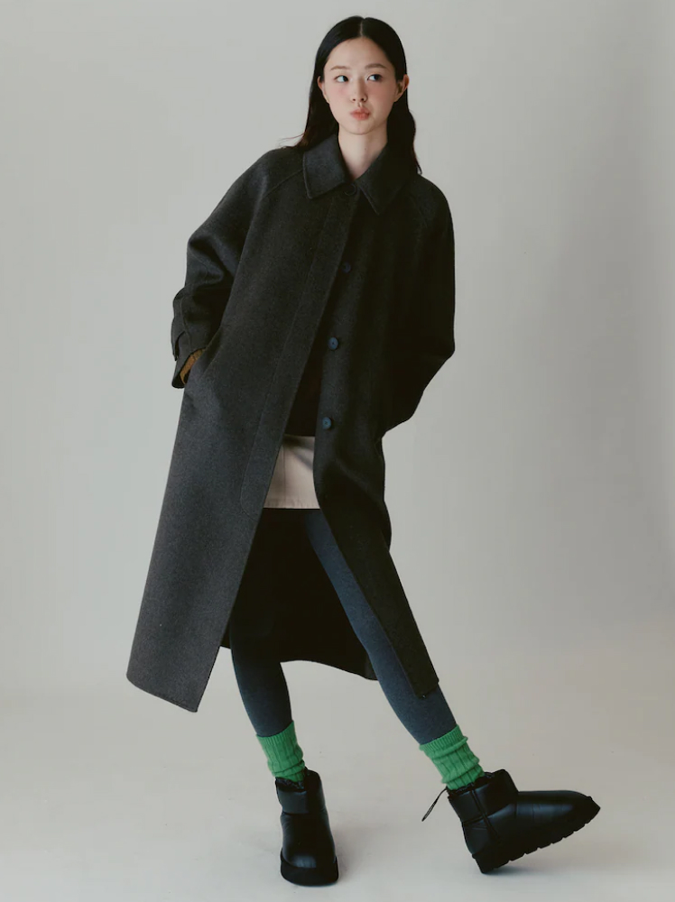 A model stares to her left side while front on with the camera and stands in a cute pose with her hip sticking out to the right while wearing a long charcoal grey coat, beige mini skirt, deep blue tights, green wool socks and a pair of black padded slipper type boots.